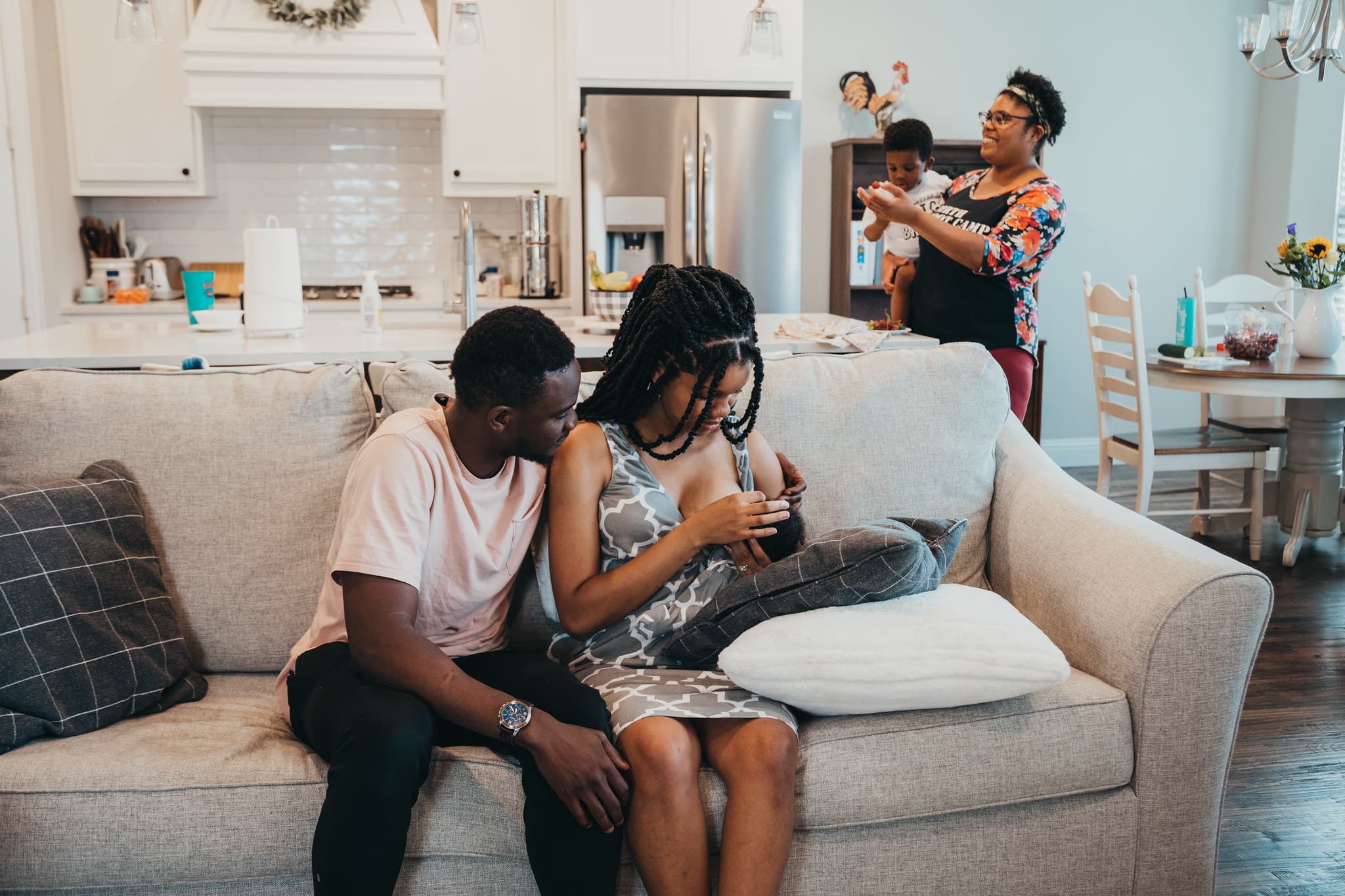 Mother breastfeeding her newborn on a couch while partner sits beside her and a postpartum doula offers support to their toddler in the background.